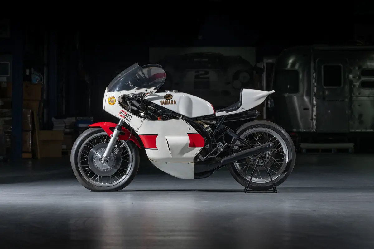 The left side profile of a 1978 Yamaha TZ750. It wears a white bubble faring at the front that wraps with the clear windscreen. The white faring wraps from the front part of the bike to panels covering the engine, with a red stripe coming over the center. There’s an inlet designed into that side panel for aero channeling. The gas tank wears white with Yamaha and it’s three tuning forks emblem in gold. The wheels are spoked wire wheels wearing slick tires
