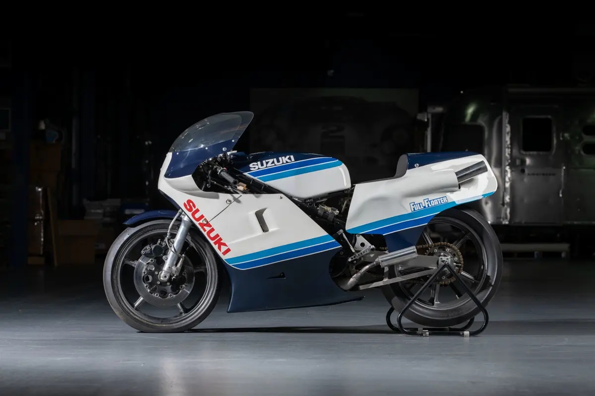 The left side of a 1983 Suzuki RGB500 Gamma wearing full carbon fiber faring painted in Suzuki coloring of a white angled striped, flanked by three stripes of blue that gradually get darker with each stripe to a navy blue taking up the bottom half or top portion of the bike. The seating is raked forward for a sport bike, and lone can only see limited details of its internal workings and mechanics due to the faring