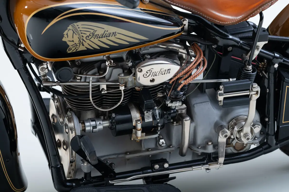A right-side close-up crop of the Indian Four model’s air-cooled, inline-four engine. A tear-drop shaped chrome piece bolted to the engine read’s ‘Indian’ in its script-like font. Here you can see the cylindrical alternator and the pedal to kick-start the engine inset