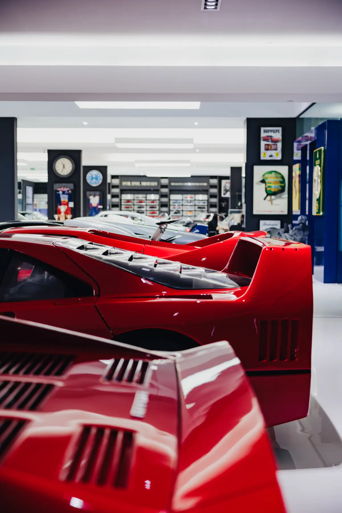 A tight vertical shot looking over the rear ends of three of the Dare to Dream Collection’s Ferraris, a Bugatti Veyron, Maserati MC12, Porsche 911… and a faint glimpse of model cars in a case at the rear of the shot on a wall. Some Porsche and other posters can be seen, along with a couple of racing suits and clocks.