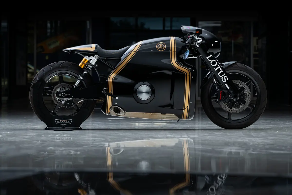 A right side profile of a 2014 Lotus C-01 wearing the John Player Special livery, on a customized Dare to Dream Garage bike stand. Here you can see a wider stance for a sportbiek, with cafe-bike-styled handlebars and end-bar, lower side mirrors, and front carbon fiber bubble faring. The black carbon fiber for the center extends from over the gas tank to under the engine like it would on a racing bike, with the center cap of the engine accessible through the faring