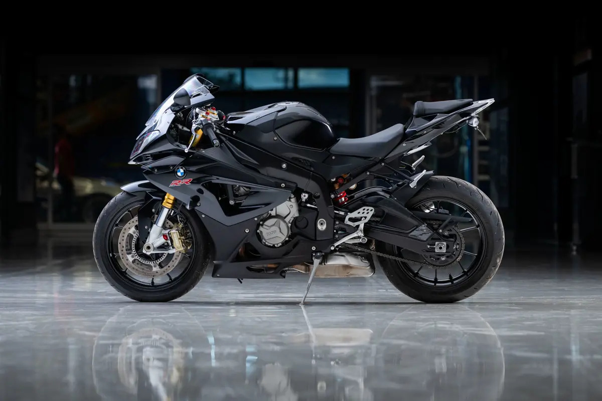 a left-side profile of a 2014 BMW S1000RR. It is an aggressive-looking bike with a shorter, sporty wheel base, racing handlebars that are cafe-style turned toward the frame instead of straight out to the sides. It is dressed in a dark grey and black carbon faring, with a front faring that angles slightly outwards towards the center of the bike. The stance and rider is raked slightly forward, as the tail end of the bike actually angles upward