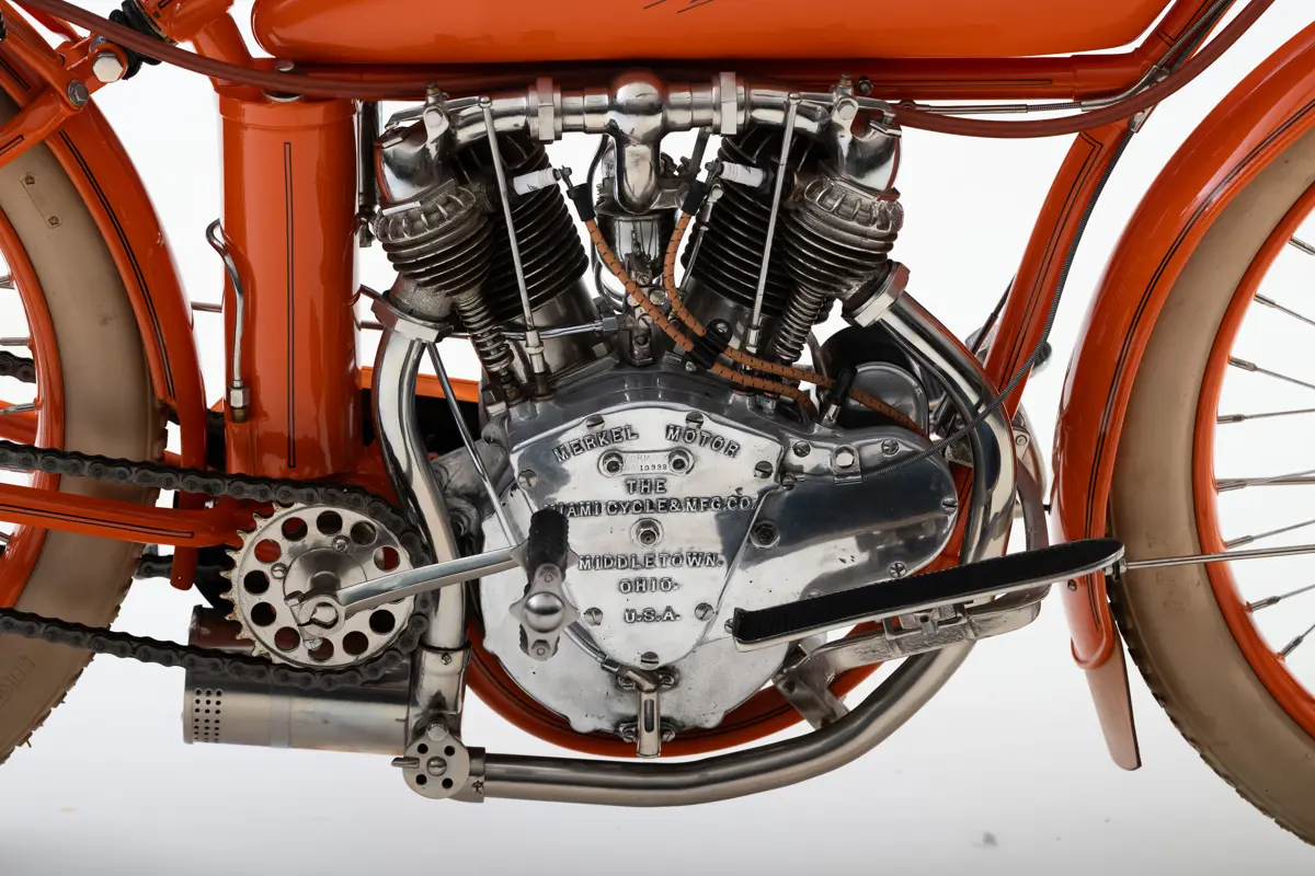 A close up of the V-twin engine with two distinctive cylinder chambers spotted with air cooling metal fins surrounding them. An upside down bell-shaped stamp is plaited to the engine reading ‘Merkel Motor. The Miami Cycle & MFG. CO. Middletown Ohio USA