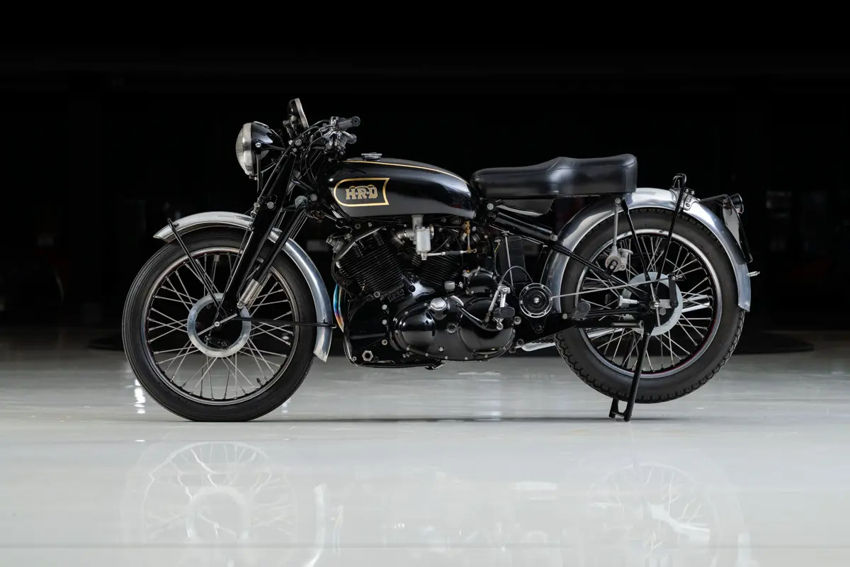 Slightly more advanced looking motorcycle of the early half of the 20th Century. This 1949 Vincent HRD Black Shadow Series C wears a black gas tank on a black frame with chrome fenders over the tires. The wheels are steel spokes, with narrow rubber tires fitted. The engine is also black, and air-cooled seen by the air-cooling fins over the cylinders